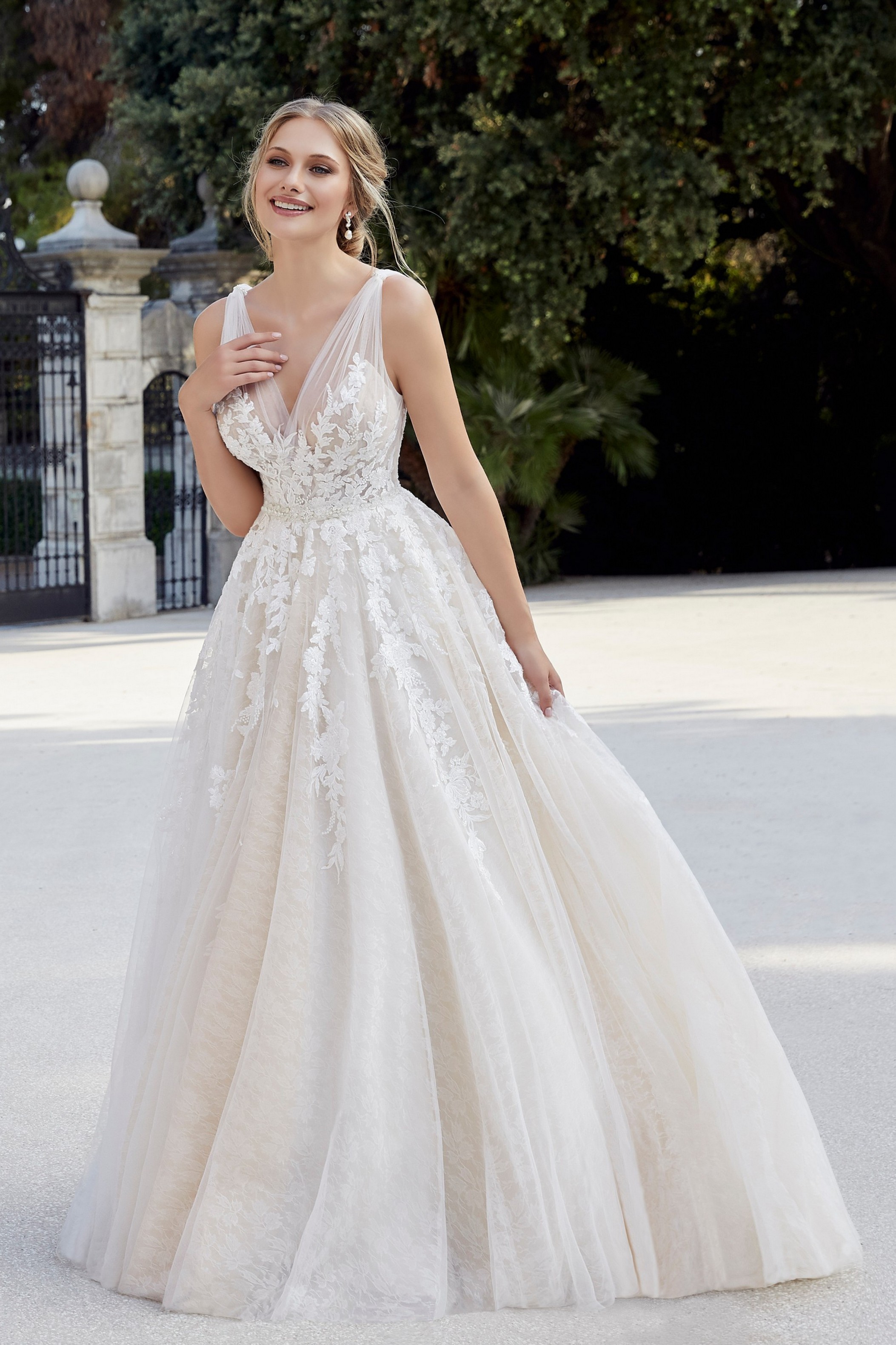 Model stood by grand black iron gates in Ronald Joyce style 18507, a vintage inspired tulle ballgown wedding dress with lace embroidery detail and ruched tulle straps
